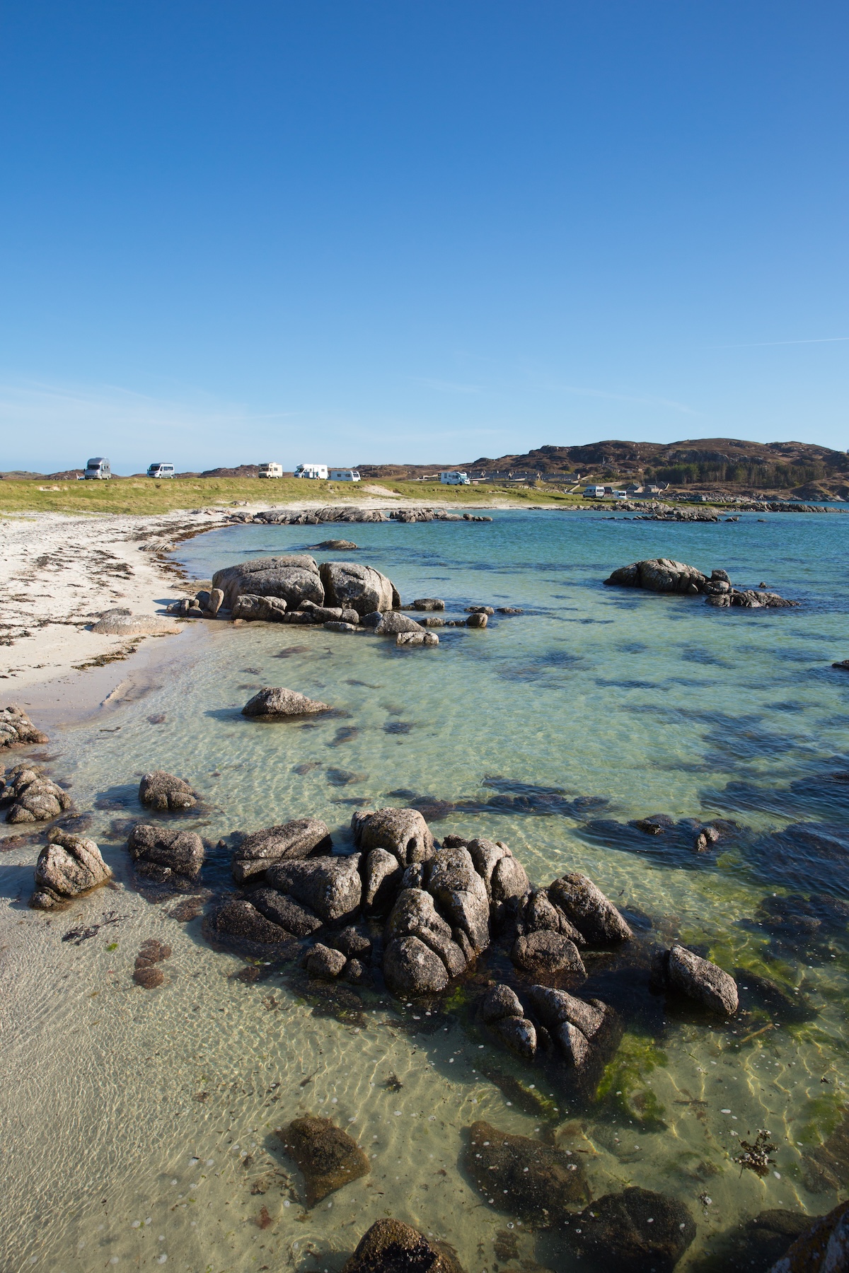 A beachy cove with a grassy bank with caravans parked along the top.